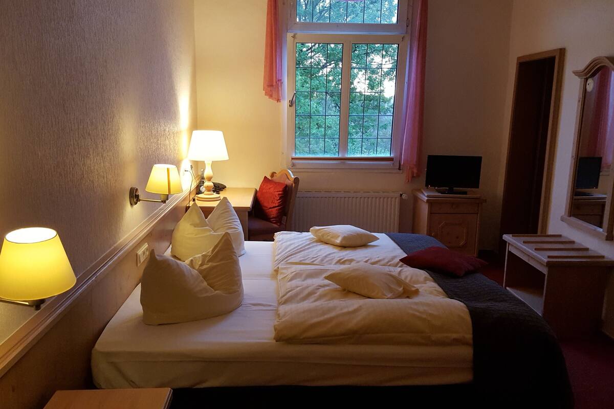 A hotel room with a bed, bedside lamps, a window with a view of trees, a desk with a computer, and a mirror.