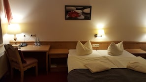 A hotel room with a bed, two bedside tables, a desk with a chair, a lamp, and a painting on the wall.