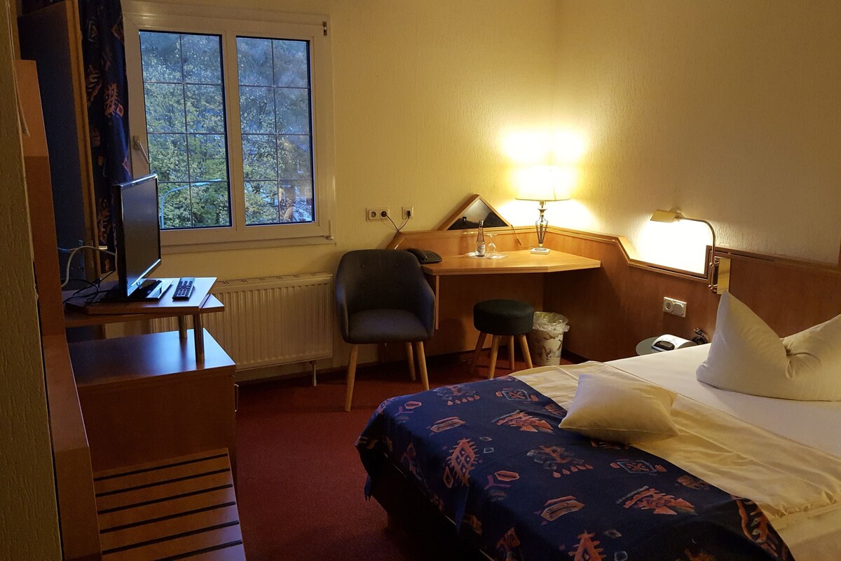 A hotel room with a bed, desk, chair, and a window with a view of trees.