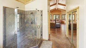 A glass-enclosed shower with stone tiles, a wooden ceiling, and a view into a living area with a sofa and a rug. - Luxury! 3 Master Suites! Located Near the Lake! Pool Table! Propane BBQ! (Big Bear City)