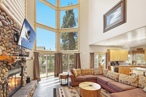 A spacious living room with a stone fireplace, large windows, a cozy sectional sofa, and a kitchen area.