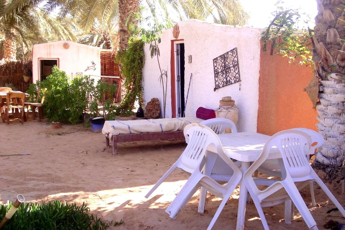 Un espace extérieur sablonneux avec une table et des chaises en plastique blanc, un banc en bois et un petit bâtiment doté d’une porte et d’une fenêtre.