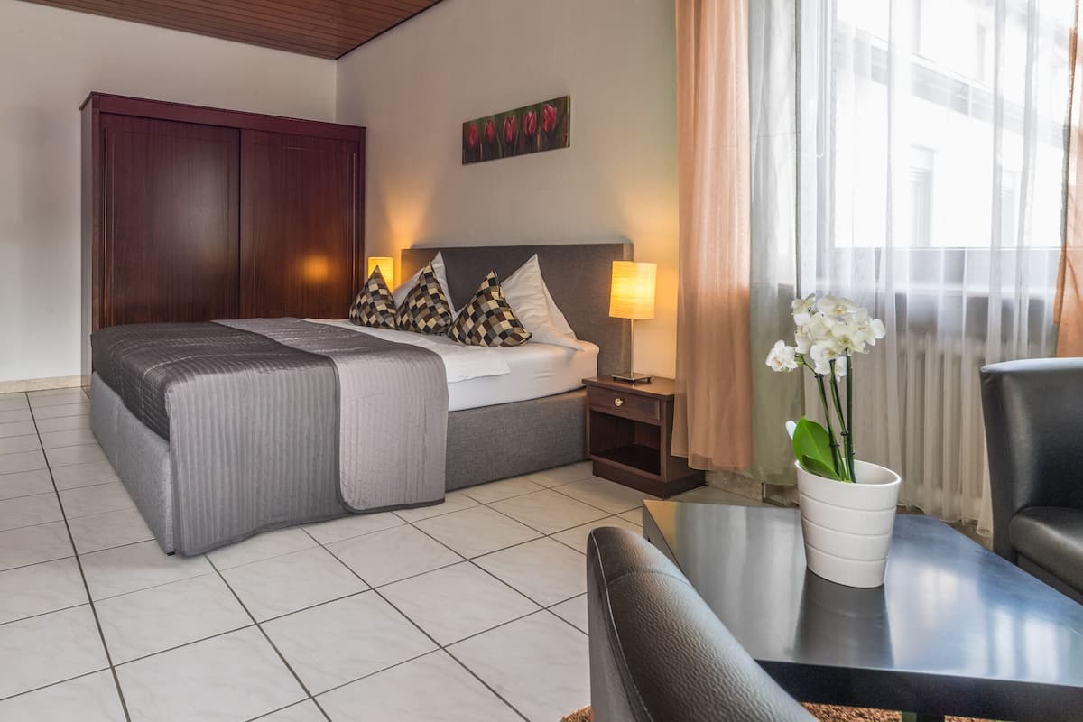 A hotel room with a bed, bedside table, lamp, chair, and a potted plant.