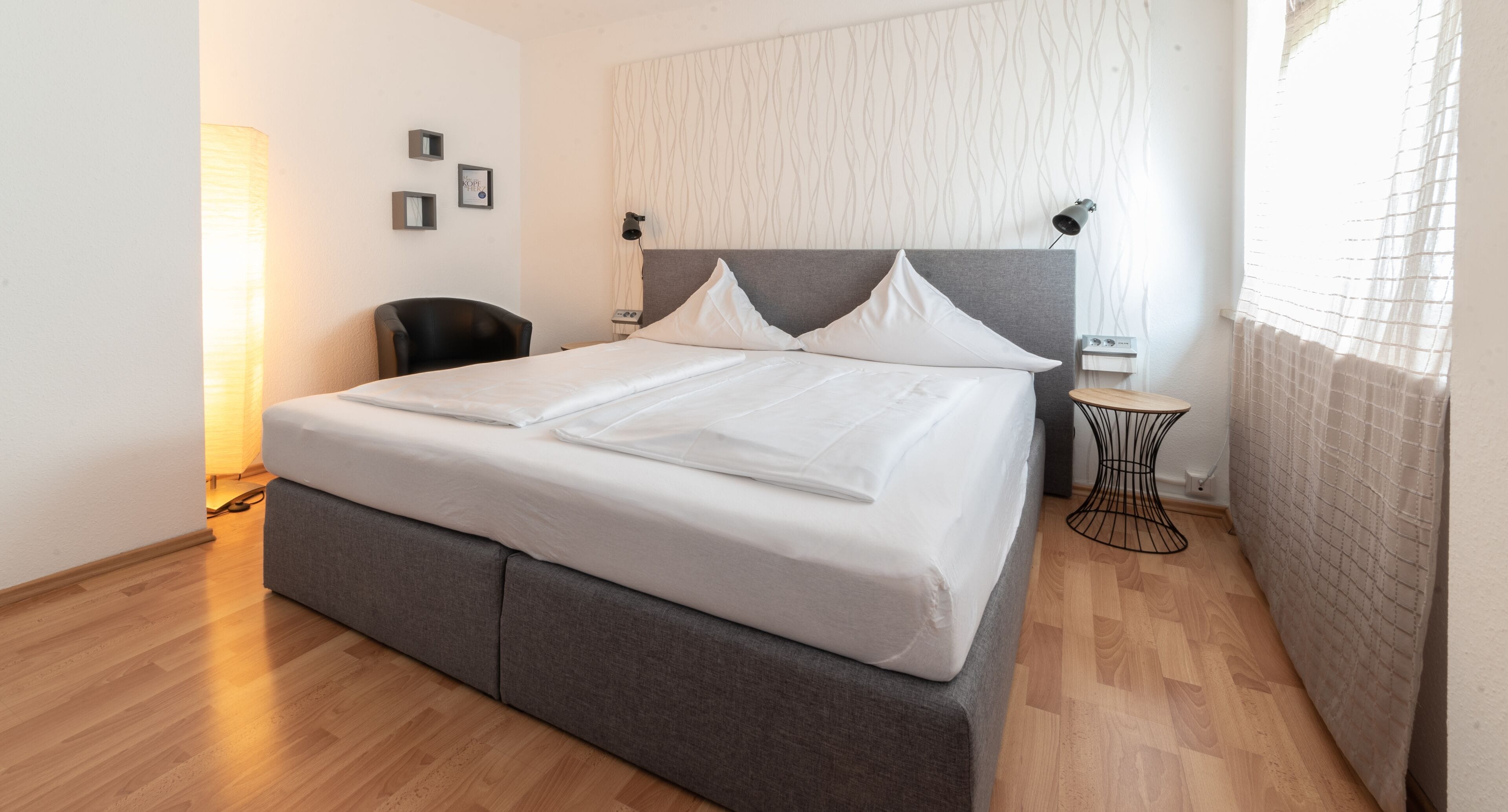 Un letto rifatto con lenzuola bianche, una testiera grigia imbottita, un comodino con una lampada e una sedia.