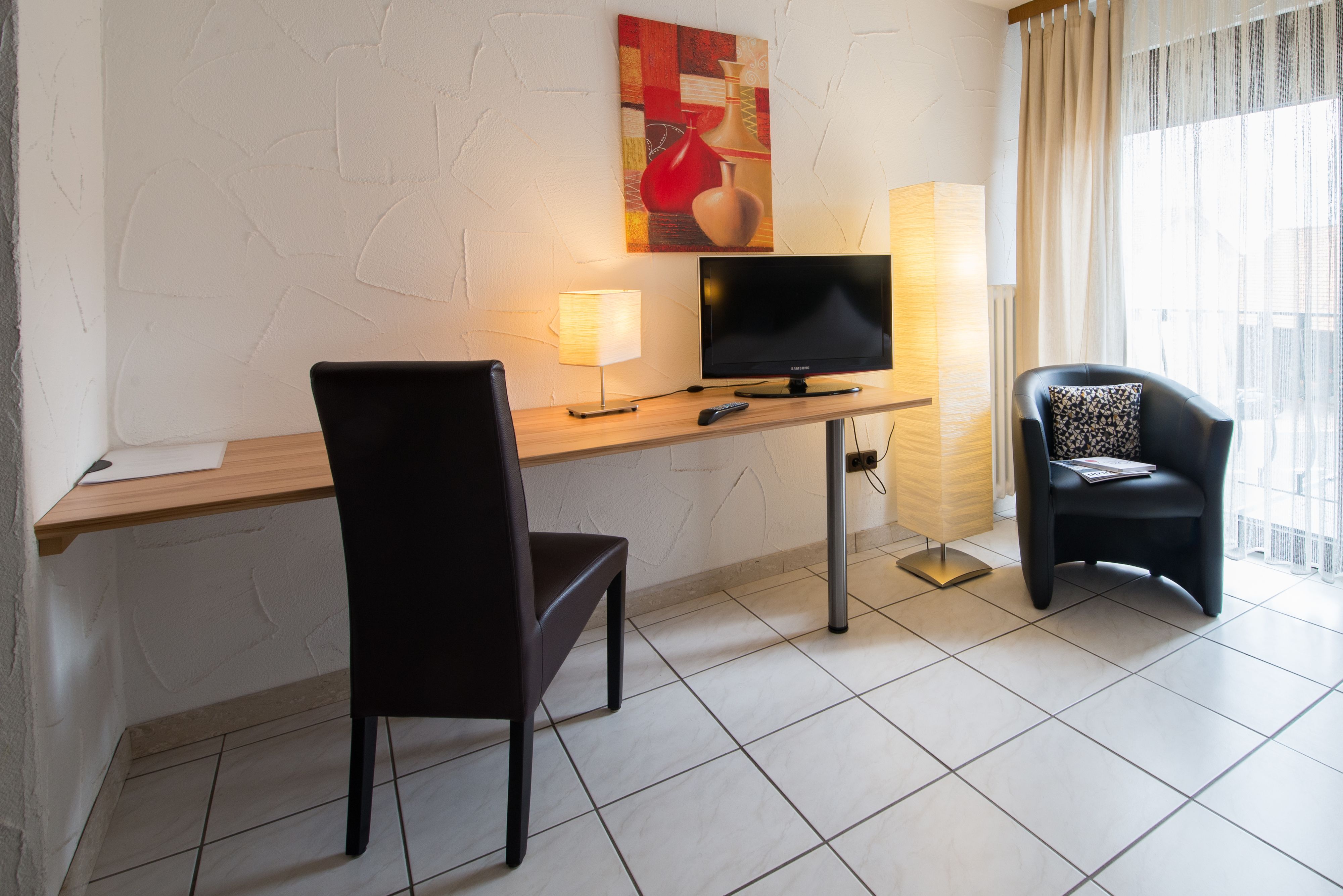 Una habitación de hotel moderna con un escritorio de madera, una silla negra, un televisor, una lámpara y una ventana con cortinas.