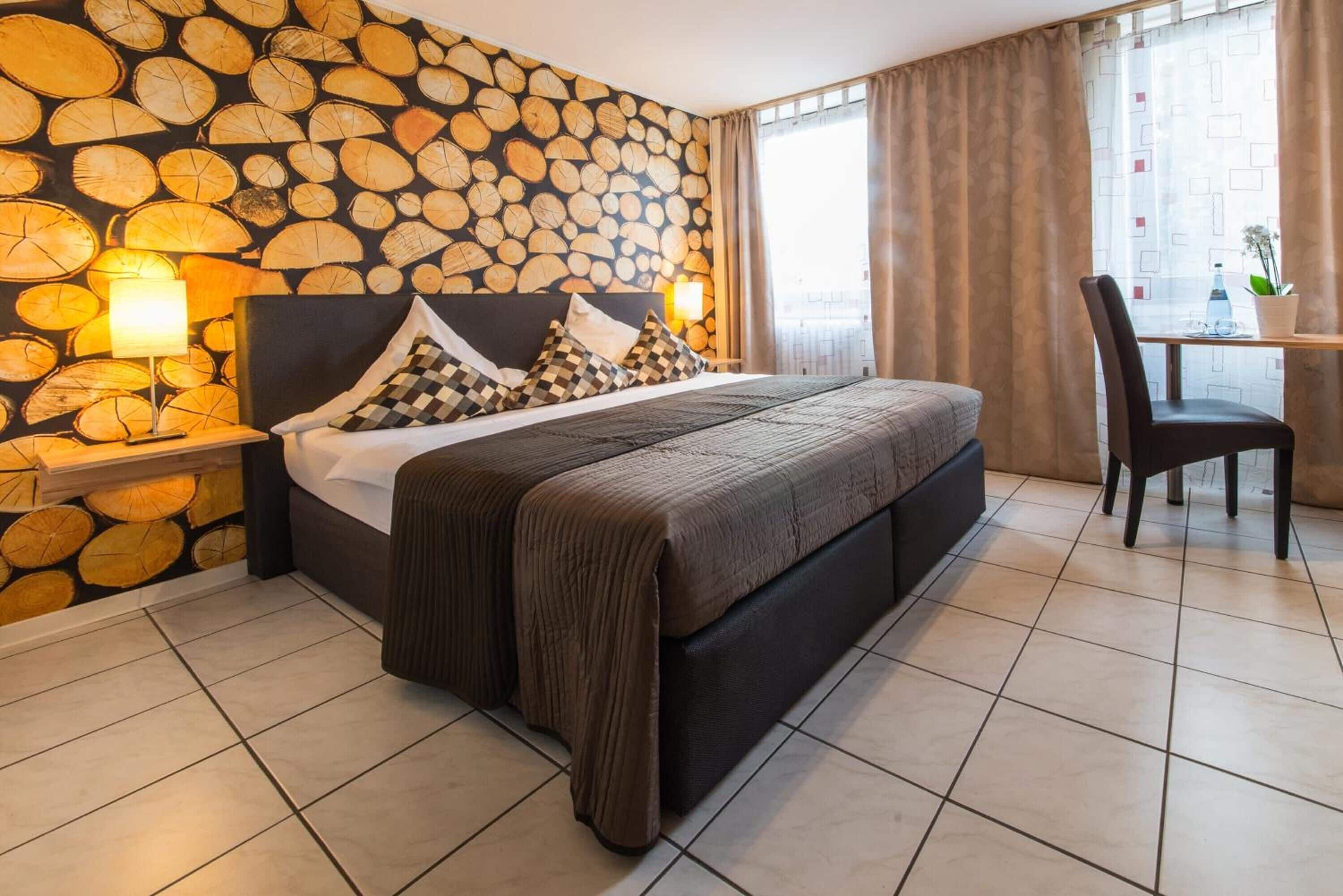 A hotel room with a bed, a nightstand with a lamp, a chair, a table with a plant, and a window with curtains.