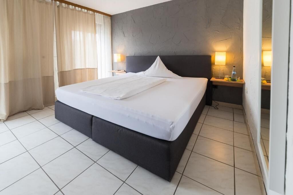 A modern hotel room with a large bed, a dark headboard, two bedside lamps, and a nightstand with a bottle and glass.
