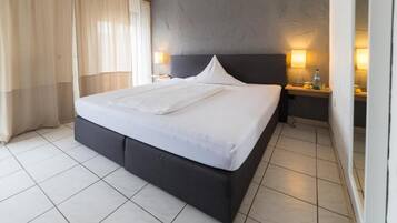 A modern hotel room with a large bed, a dark headboard, two bedside lamps, and a nightstand with a bottle and glass.