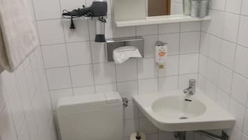 Un baño pequeño y moderno con inodoro, lavamanos y espejo. Se observa un toallero, un estante pequeño con una planta y un estante de madera con una toalla enrollada.