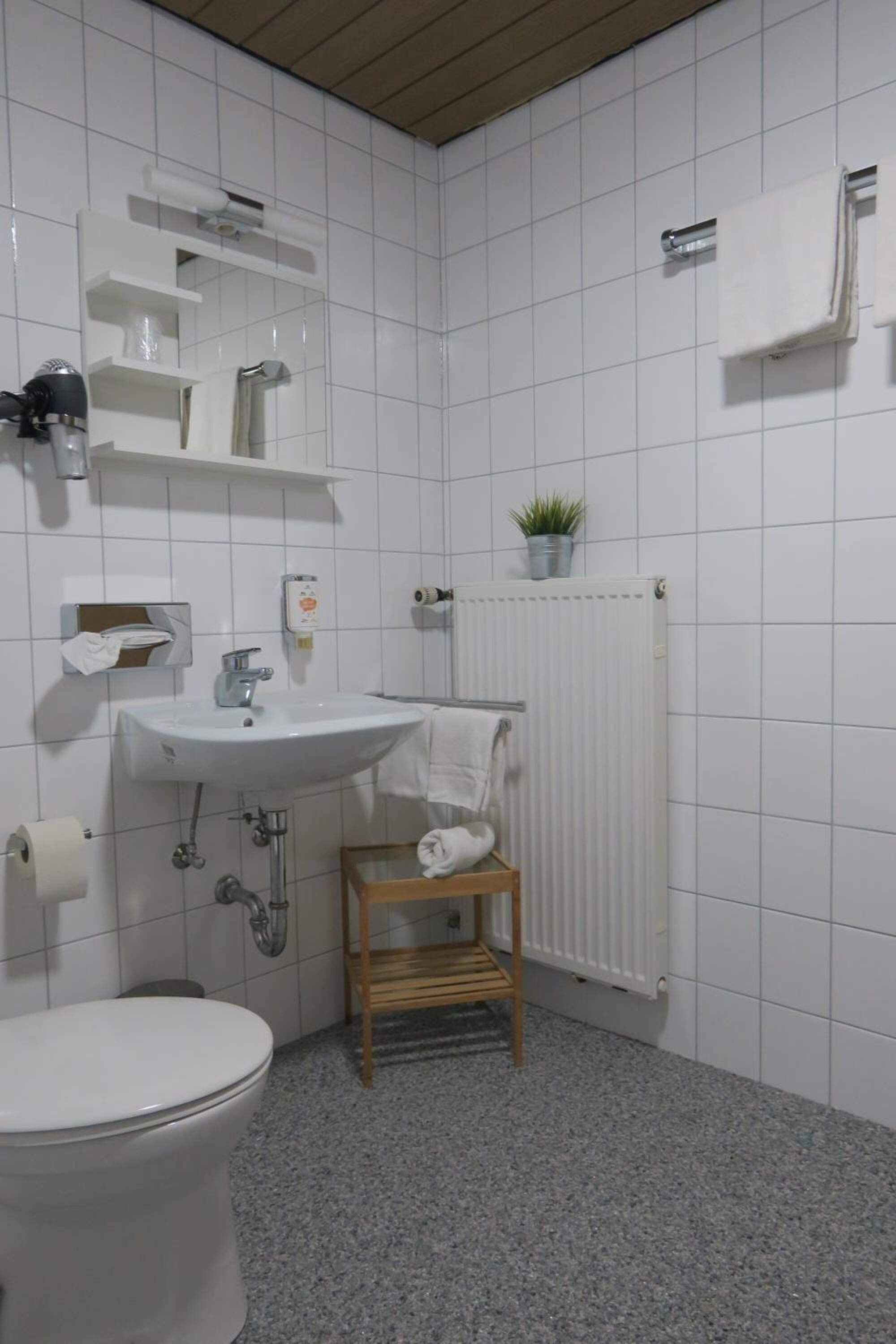 A bathroom with a white sink, toilet, and a small wooden stool. There is a towel rack with a towel hanging on it, a white radiator, and a potted plant on the radiator.