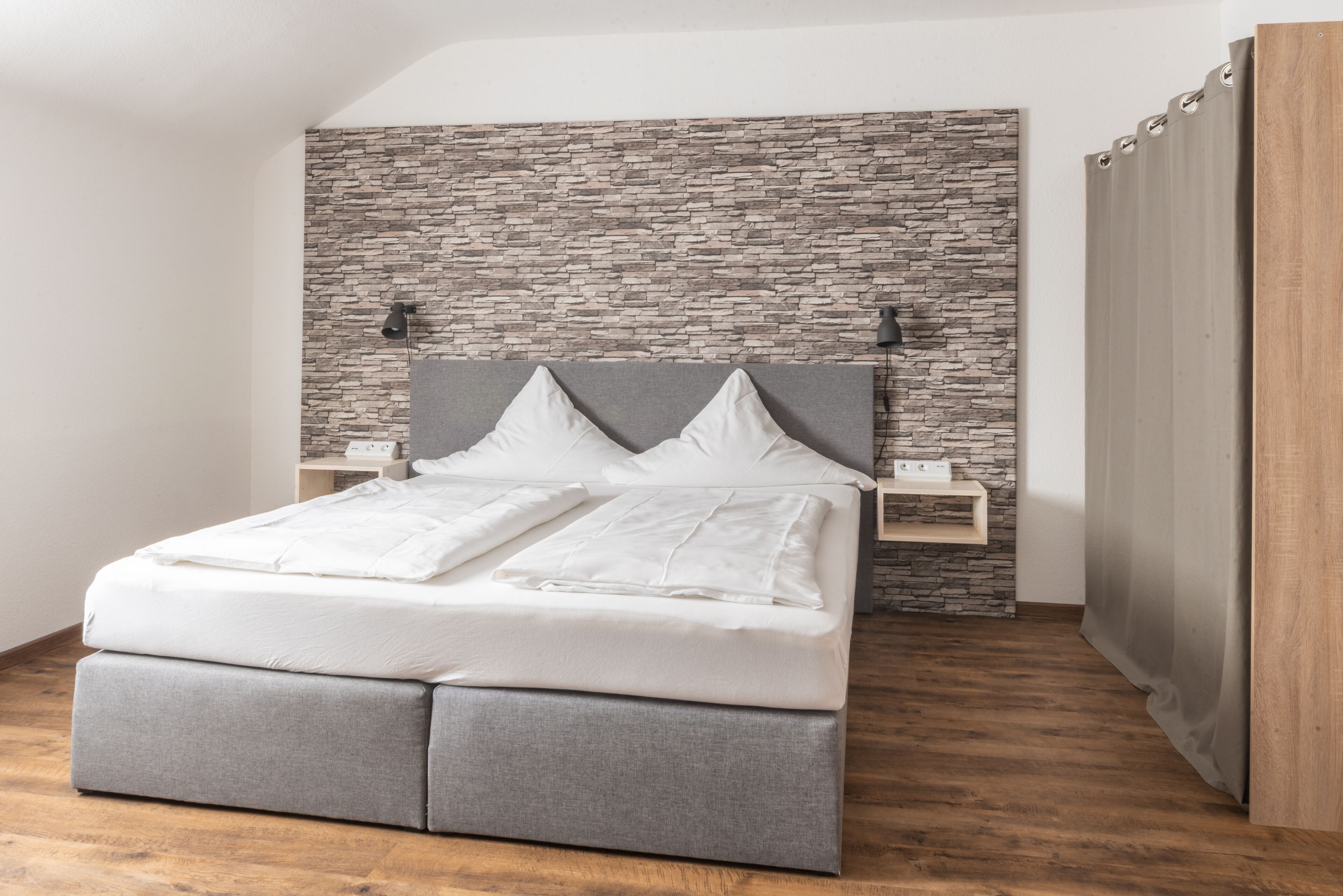 Une chambre moderne avec une tête de lit en pierre, un lit recouvert d’un tissu gris, une table de chevet avec une lampe et un plancher en bois.