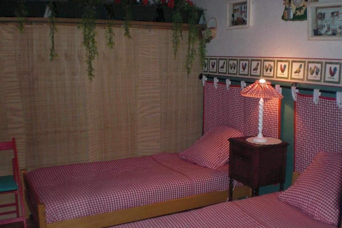Ein Schlafzimmer mit zwei Einzelbetten, einem Nachttisch mit Lampe, einem Sessel und einem Holz-Kopfteil mit Wanddekorationen.