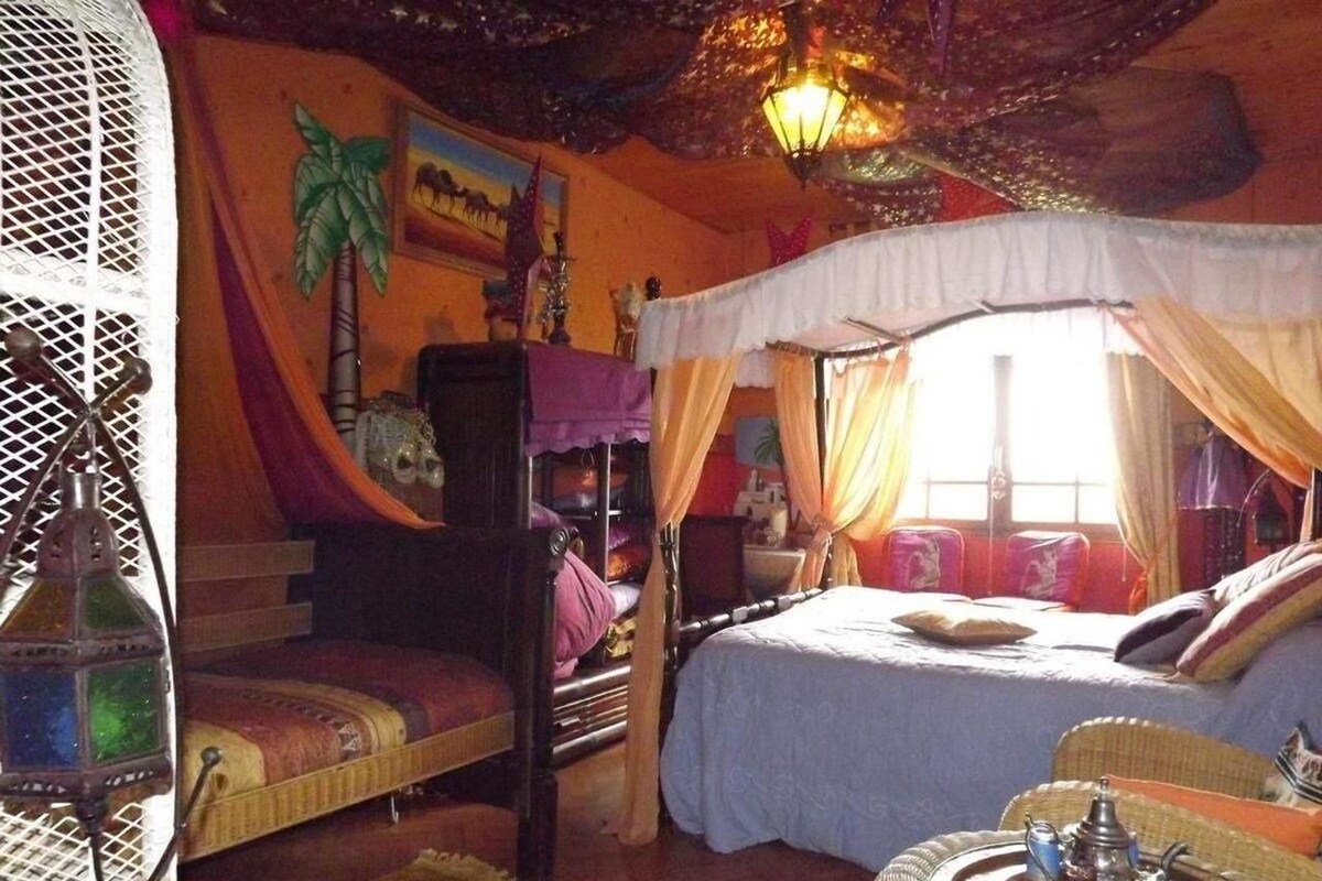 Ein Zimmer mit einem Bett unter einem Baldachin, einem kleinen Tisch mit Teekanne und einer Wand mit einem Palmenbaumgemälde.