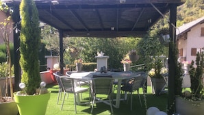 Une terrasse extérieure couverte, dotée d’une table ronde et de chaises, entourée de plantes en pot et d’une pelouse.