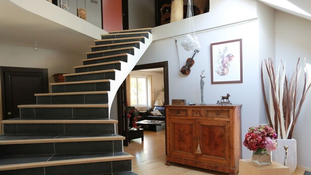 Un escalier aux carreaux sombres et aux marches en bois mène au deuxième étage. On aperçoit, au premier plan, une armoire en bois dotée de tiroirs et un vase rempli de fleurs.