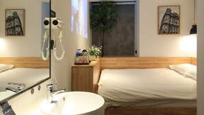 Une chambre d’hôtel avec un lit en bois, un lavabo rond blanc, un miroir, une petite table de chevet avec une plante, et deux tableaux encadrés au mur.