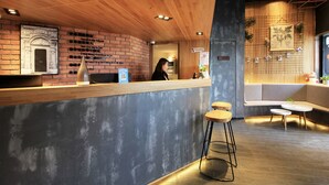 Un espace d’accueil moderne avec un comptoir en bois, un coin salon avec un canapé et des tables, et un mur en briques agrémenté d’une arche décorative.