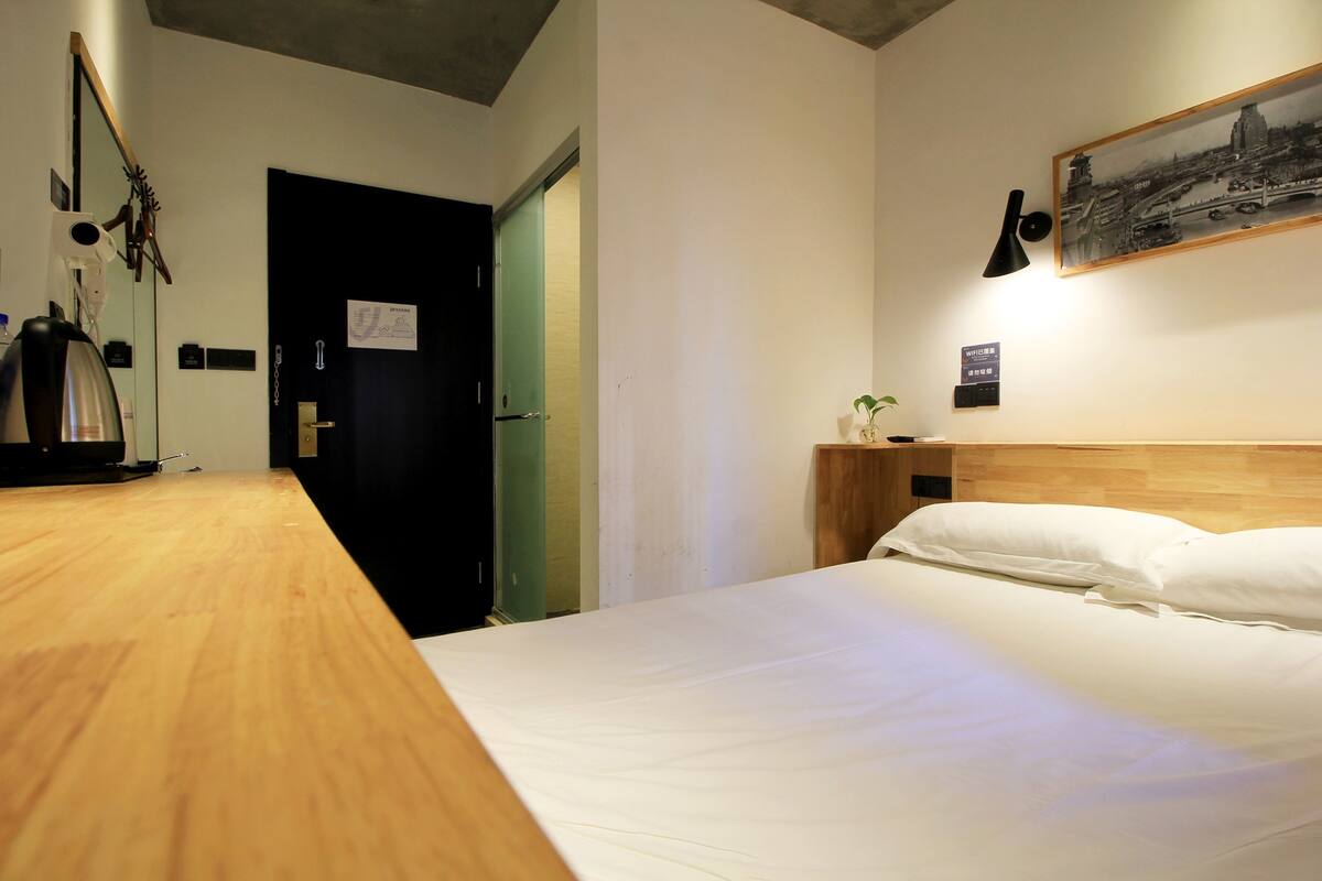 Een hotelkamer met een houten bureau, een bed met witte lakens, een nachtkastje met een lamp en een ingelijst schilderij aan de muur.