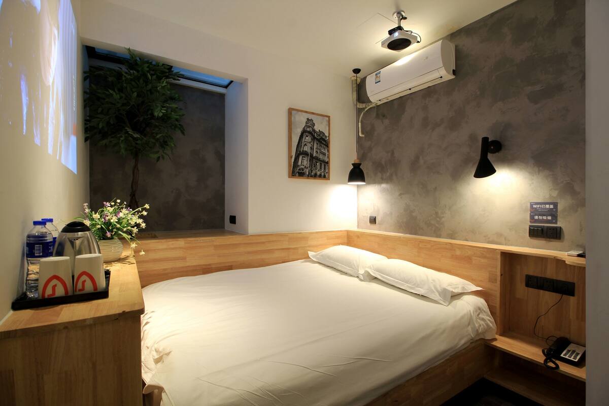 Een moderne hotelkamer met een houten hoofdeinde, een bed met witte lakens, een klein nachtkastje met een lamp, een wandairconditioner en een ingelijst schilderij aan de muur.