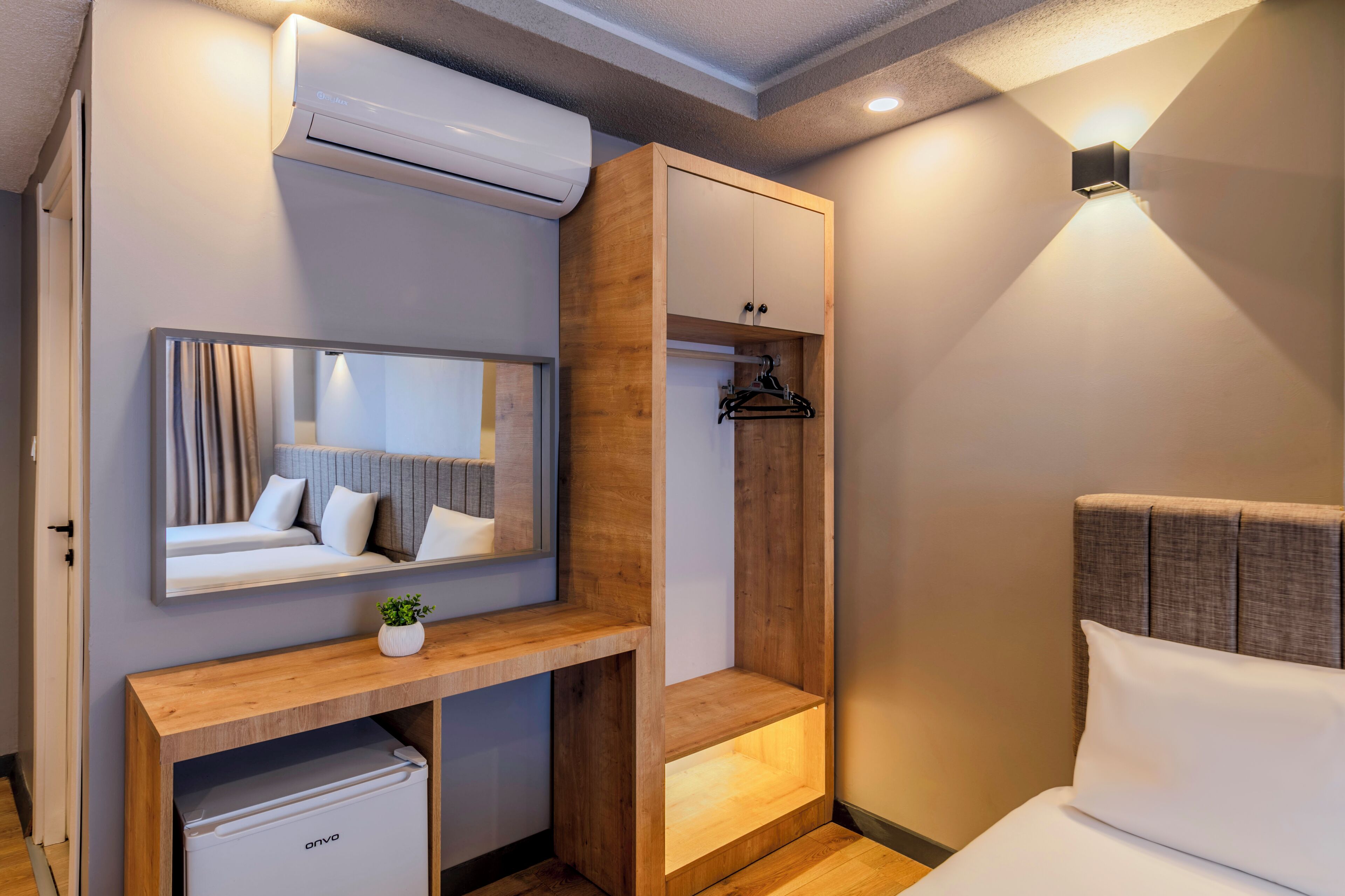 A modern bedroom with a built-in wardrobe, a bedside table, a small fridge, a mirror, and a wall-mounted light fixture.