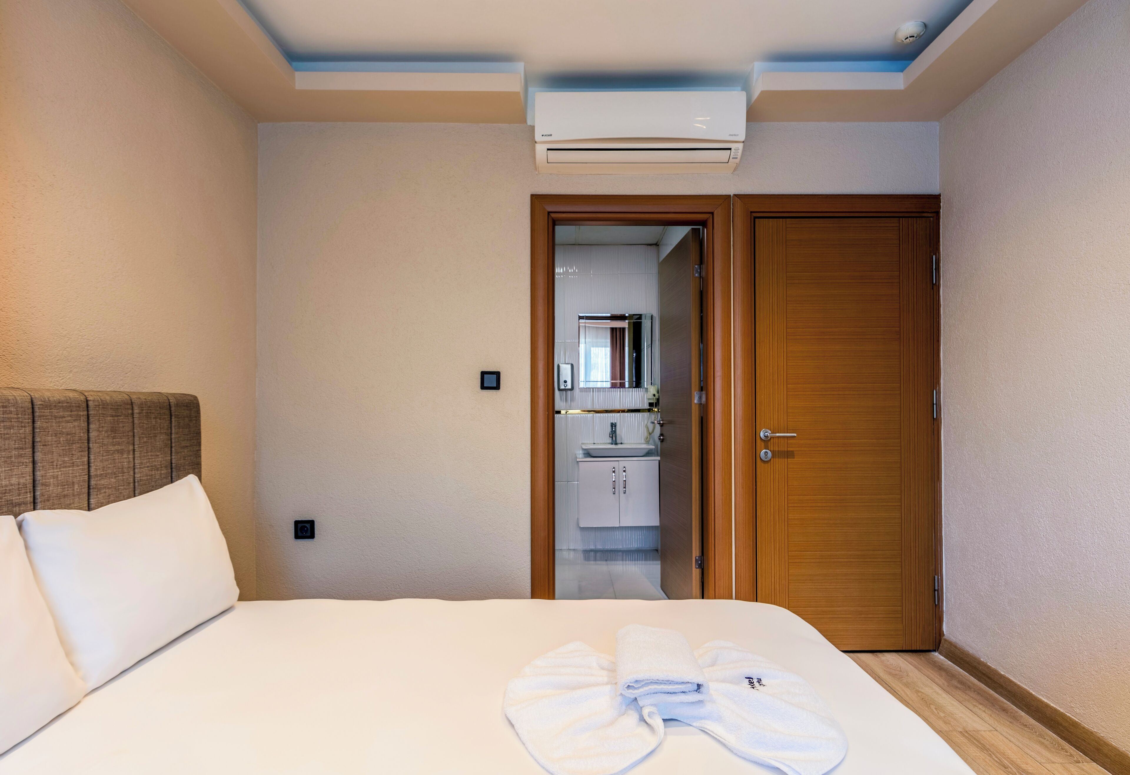 A hotel room with a bed, a wooden door, an air conditioning unit, and a bathroom visible through an open door.