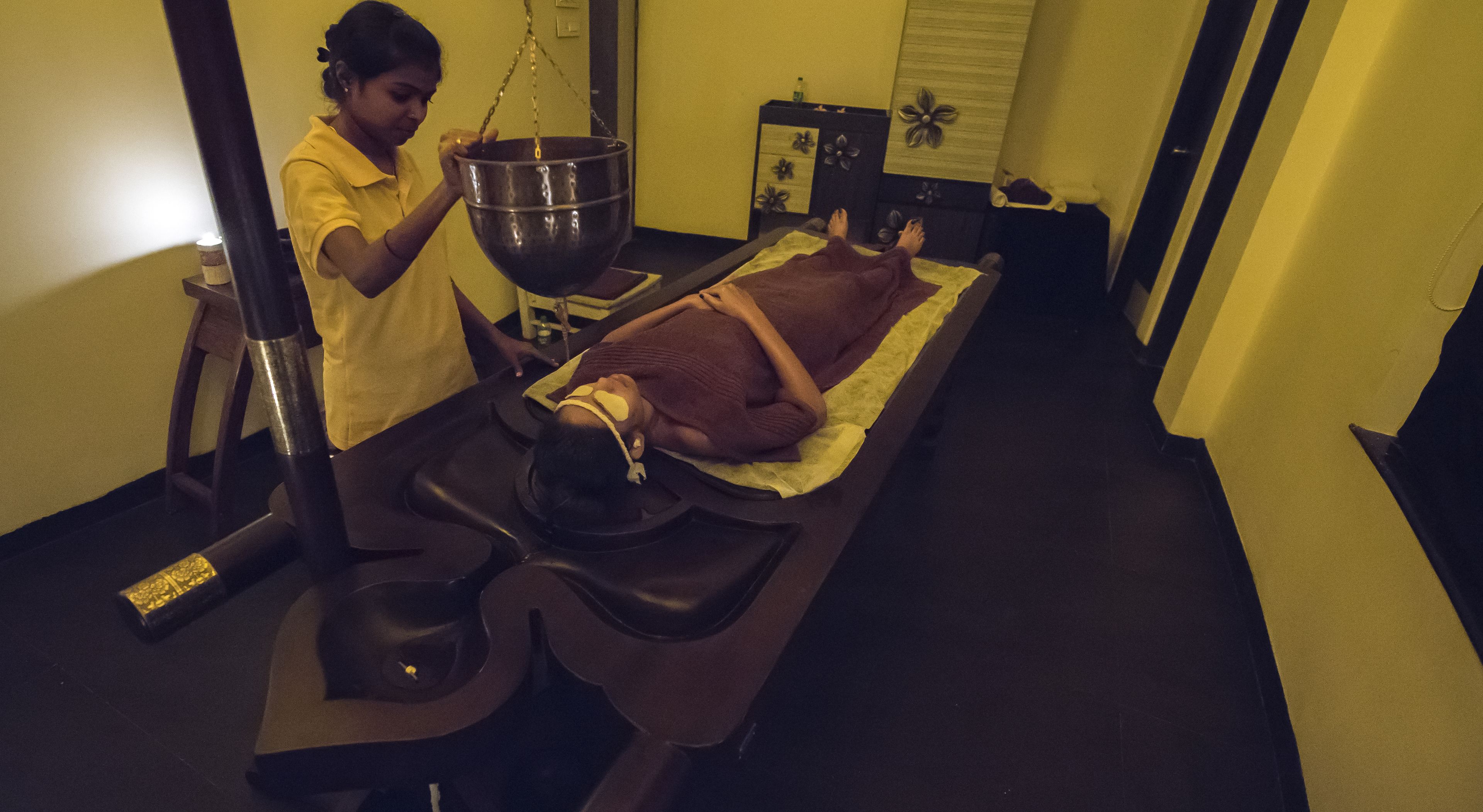 A massage therapy room with a massage table, a large metal bowl, and a person performing a massage.