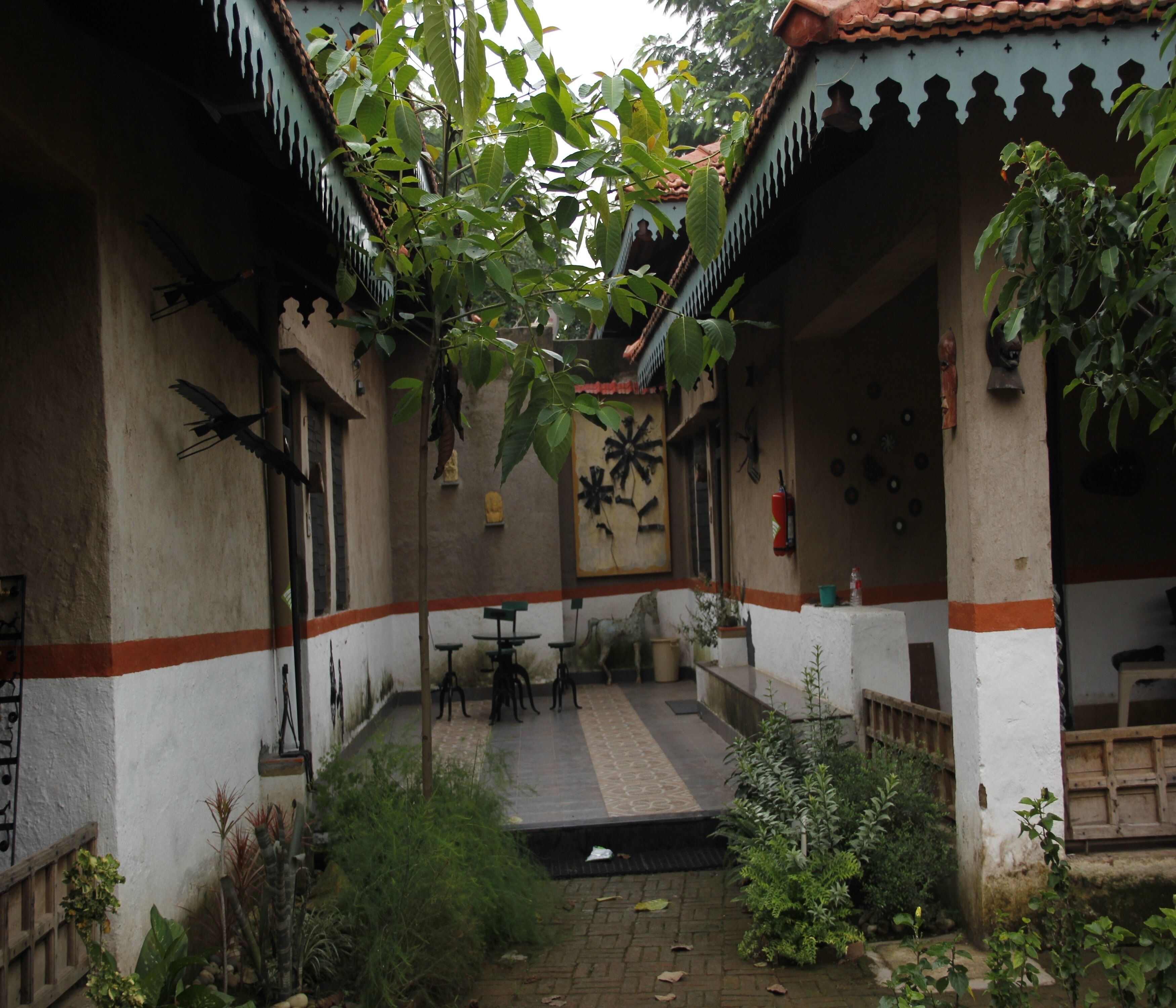 A courtyard with a tiled floor, surrounded by walls with decorative patterns and a tiled roof. There are tables and chairs in the center, and various plants and trees around the perimeter.