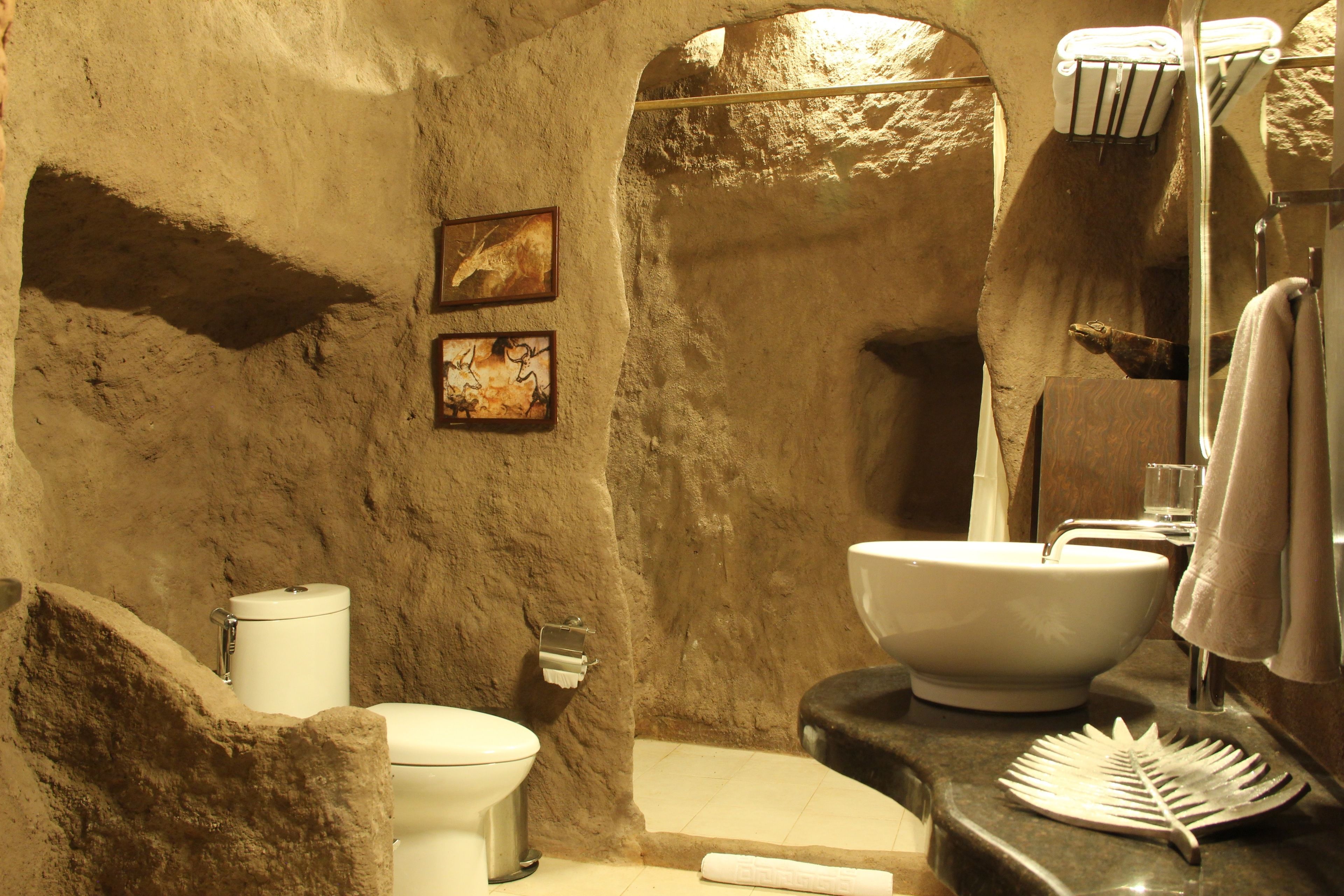 Un baño con pared de piedra rústica, un inodoro blanco, un lavamanos con un jabonera con forma de hoja y una toalla colgada en la pared.