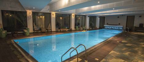 An indoor swimming pool with clear blue water, surrounded by wooden flooring and large windows.