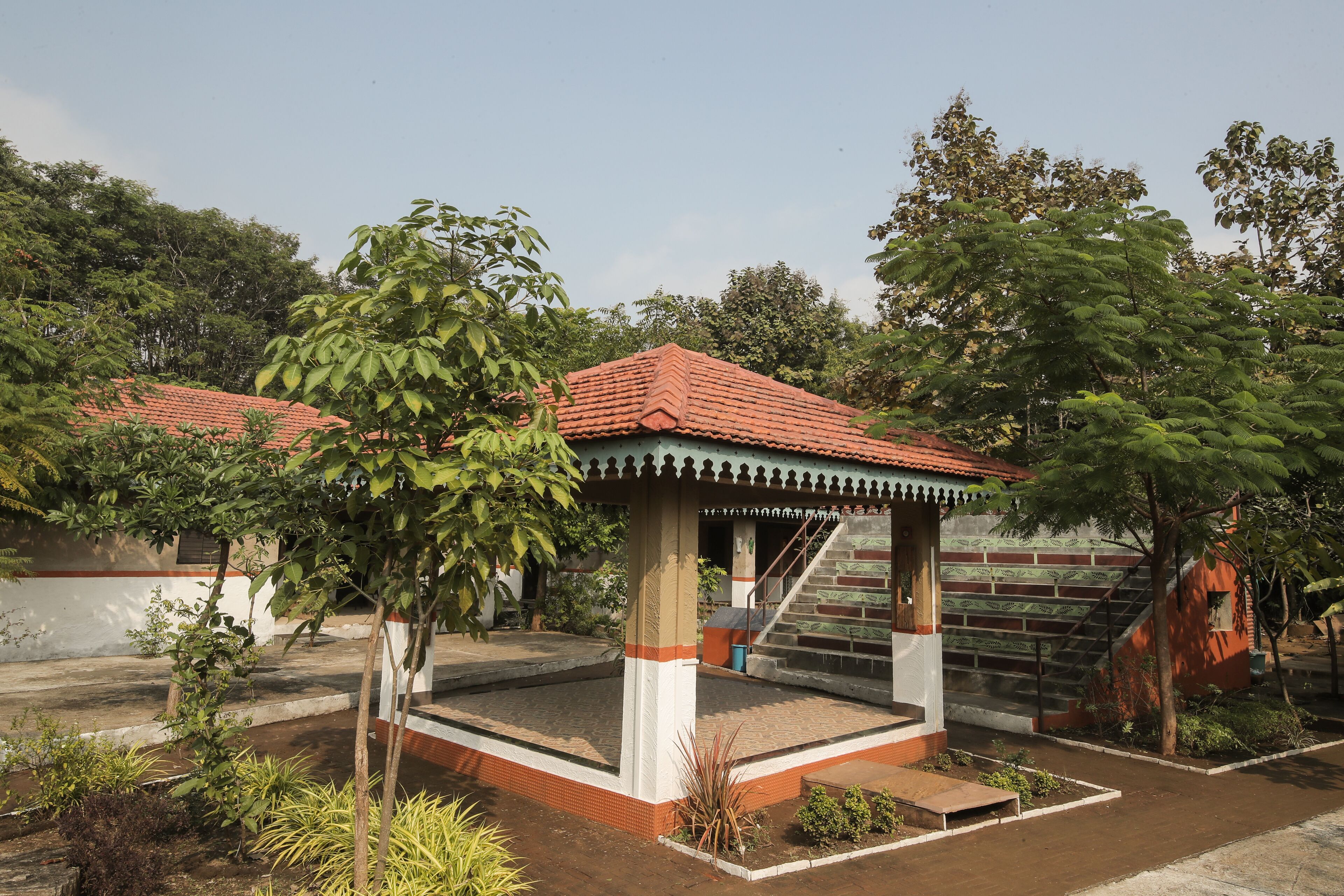 A gazebo with a tiled roof, surrounded by greenery and a staircase leading to an upper level.
