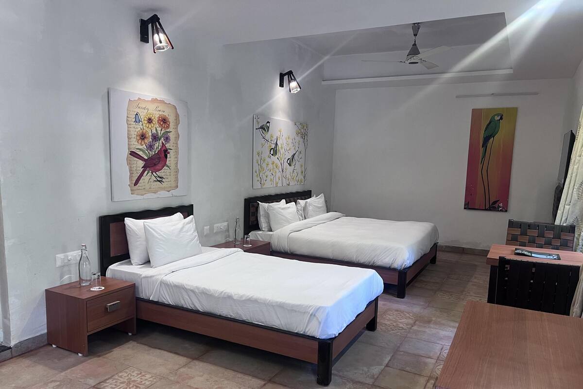 A hotel room with two single beds, a wooden desk, and a chair. There are two bedside tables with lamps, a ceiling fan, and artwork on the walls.