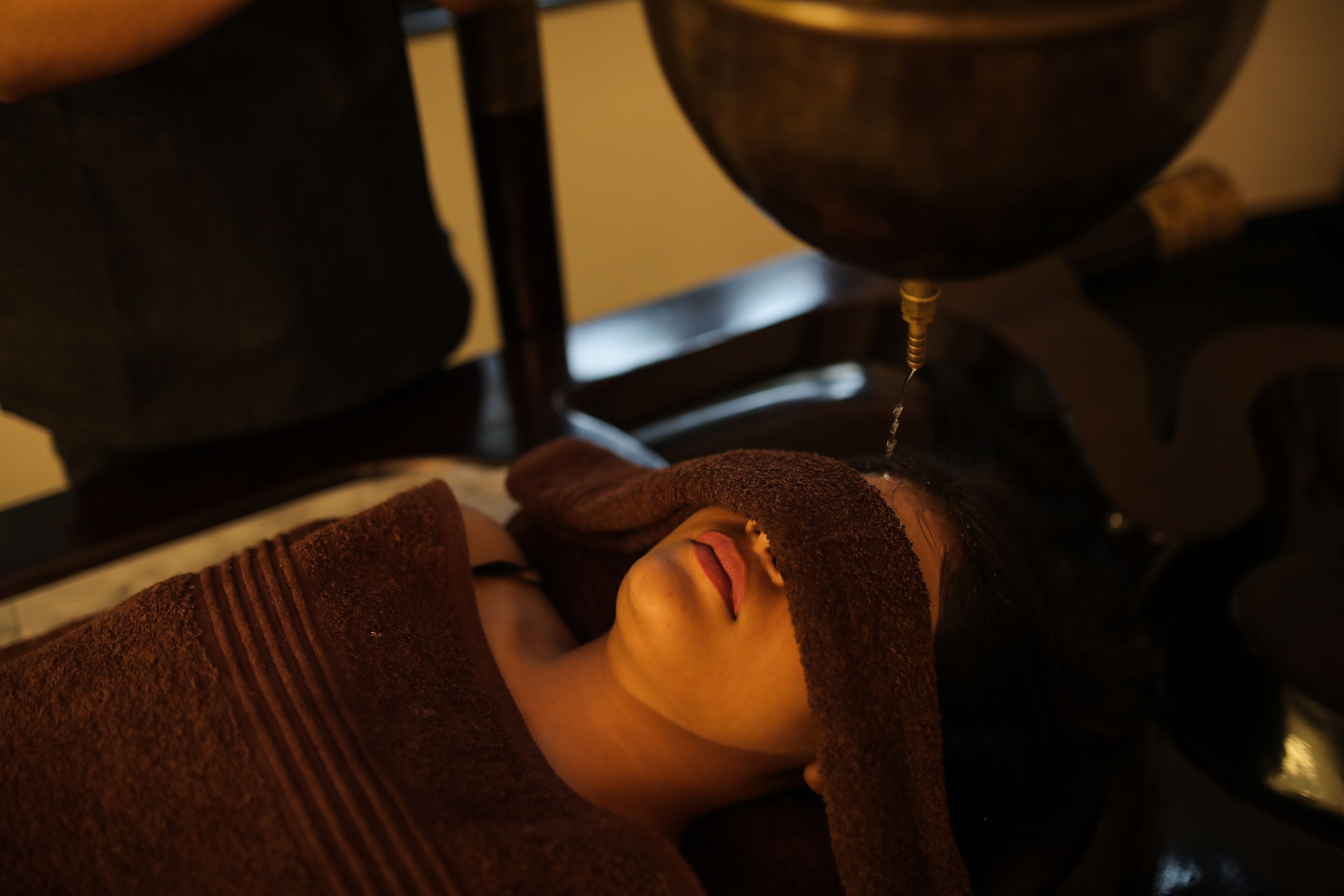 A person lying down with a towel wrapped around their head, receiving a treatment from a metal bowl.