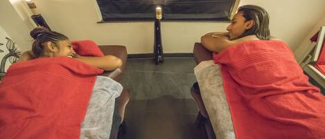 Two people sitting on chairs with red towels, a candle on a stand, and a window with a curtain.