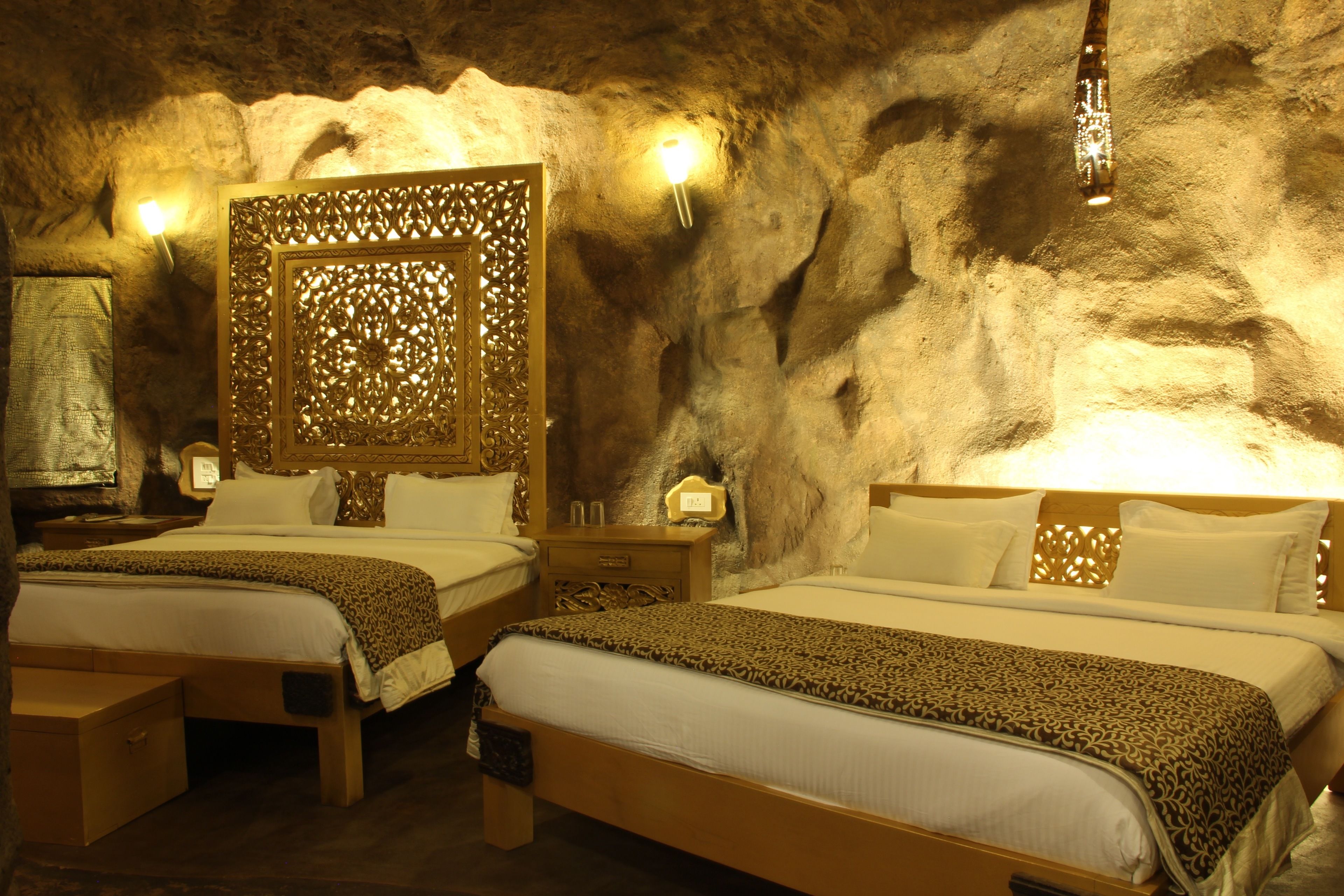 A room with two beds, each with a wooden frame and white bedding, set against a textured wall with a decorative panel and wall-mounted lights.