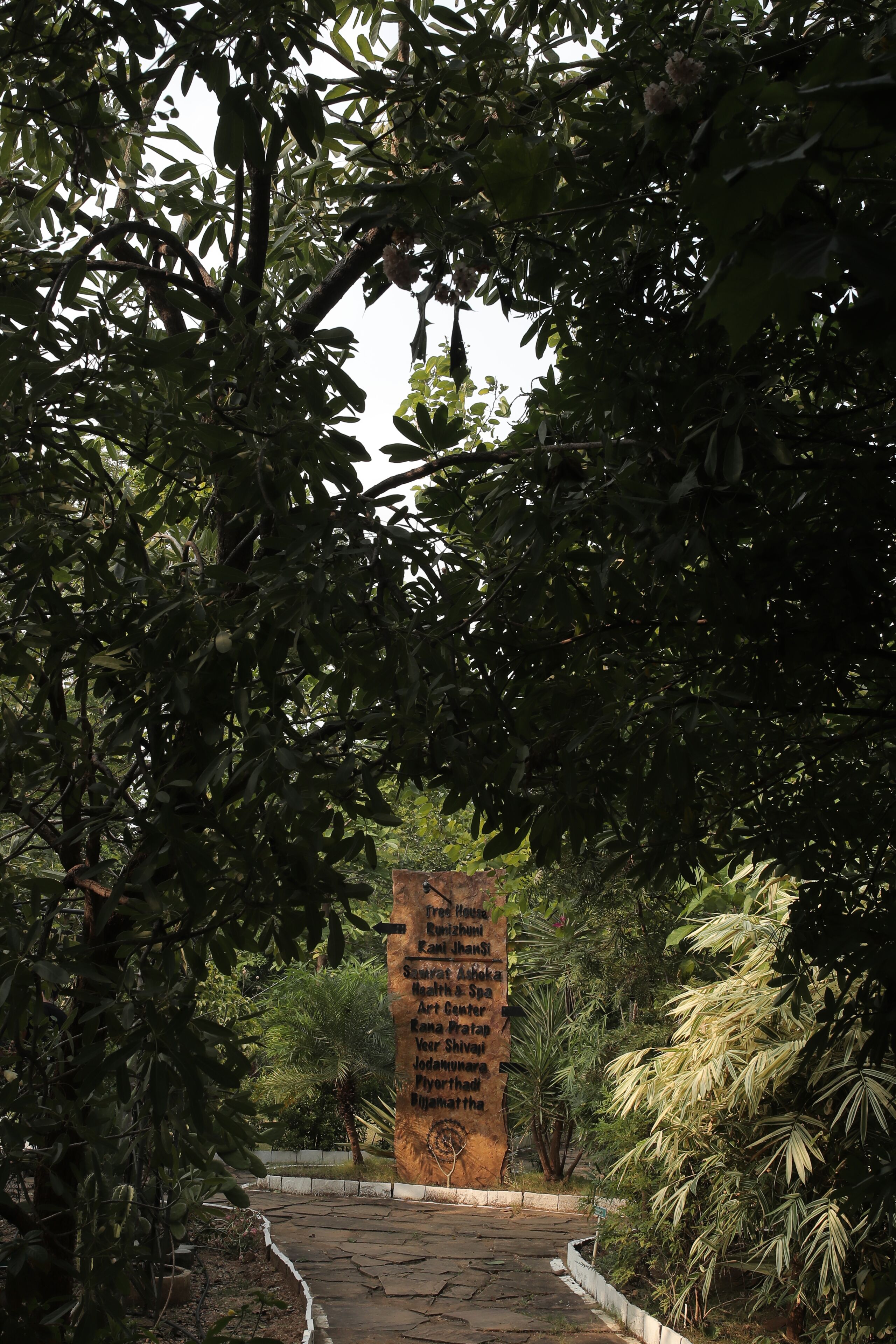A stone pathway leading to a central pillar with inscriptions, surrounded by lush greenery and trees.