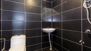 A small, dark-tiled bathroom with a white toilet, a pedestal sink, and a showerhead.