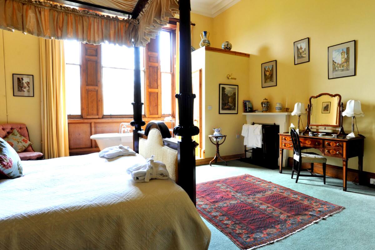 Un lit à baldaquin, un tapis aux motifs rouges et bleus, un bureau en bois avec une chaise, et une commode avec un miroir et des lampes.
