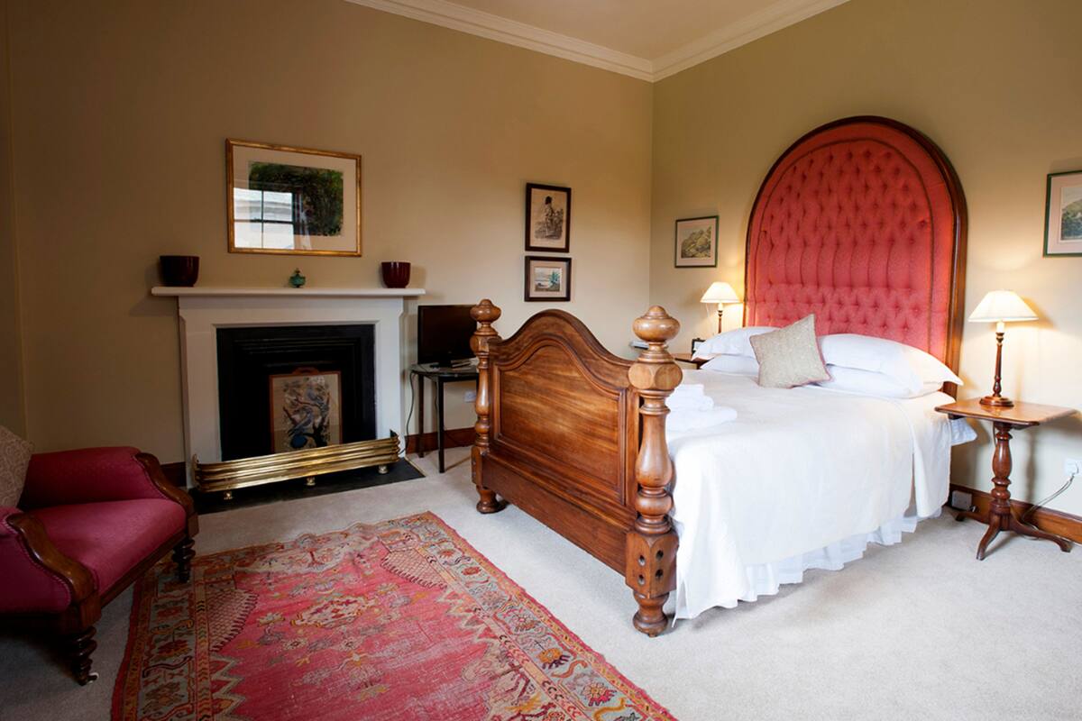 Une chambre avec un grand lit, une cheminée, un fauteuil rouge et un tapis à motifs.