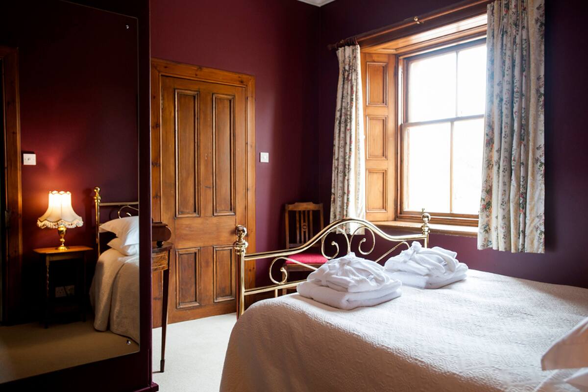 Une chambre d’hôtel avec un lit en bois, une table de chevet avec une lampe, une armoire et une fenêtre ornée de rideaux à motifs floraux.