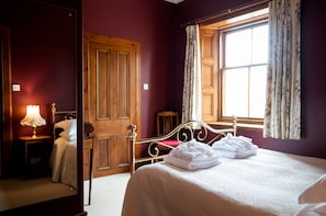 A hotel room with a wooden bed, a nightstand with a lamp, a wardrobe, and a window with floral curtains. - Cambo House And Estate B&B (St. Andrews)