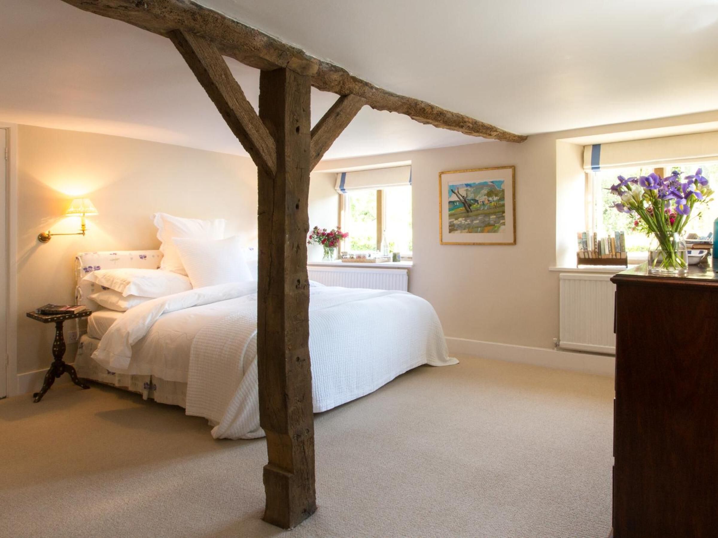 A bedroom with a large bed, a nightstand, a lamp, a window with a view, and a wooden beam structure.
