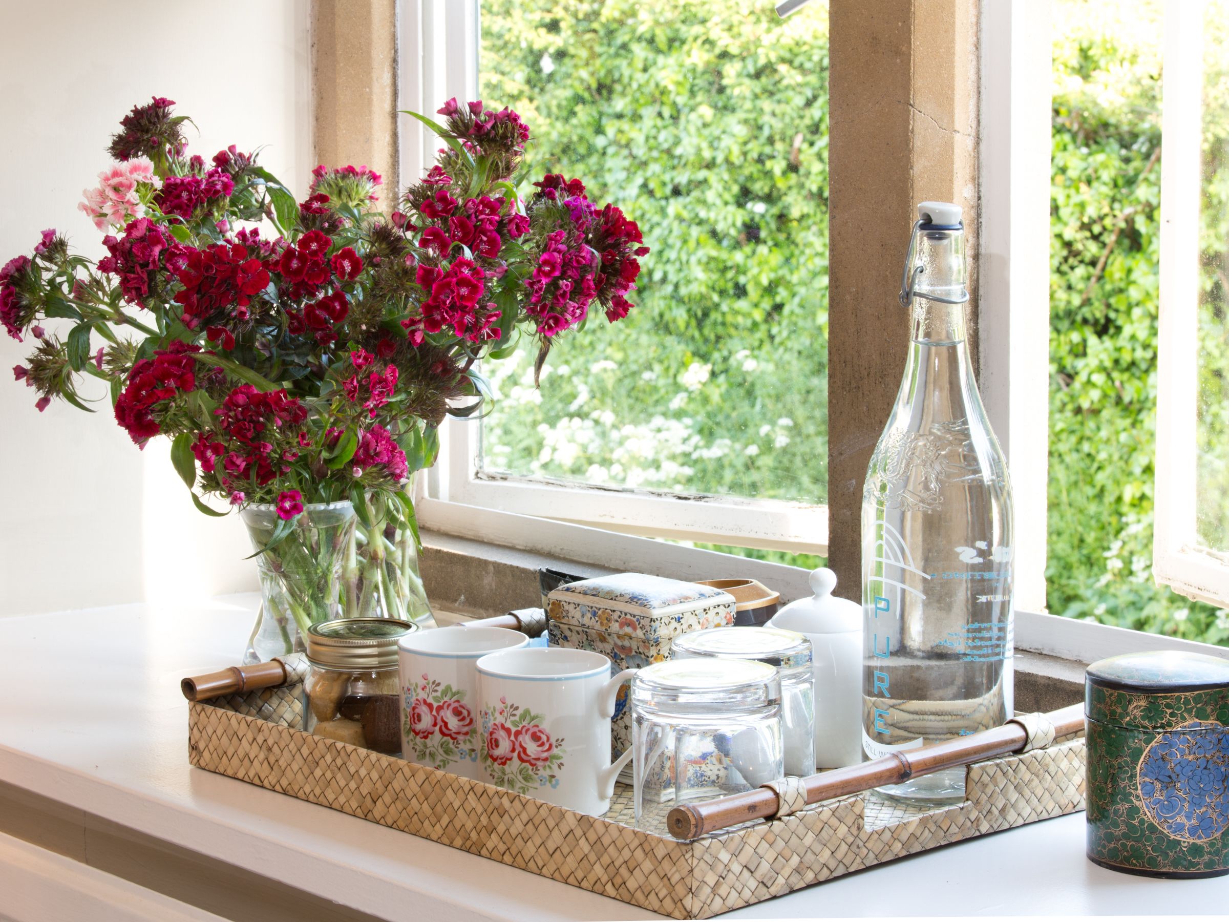 A tray with a vase of flowers, a bottle, and various containers on a windowsill.
