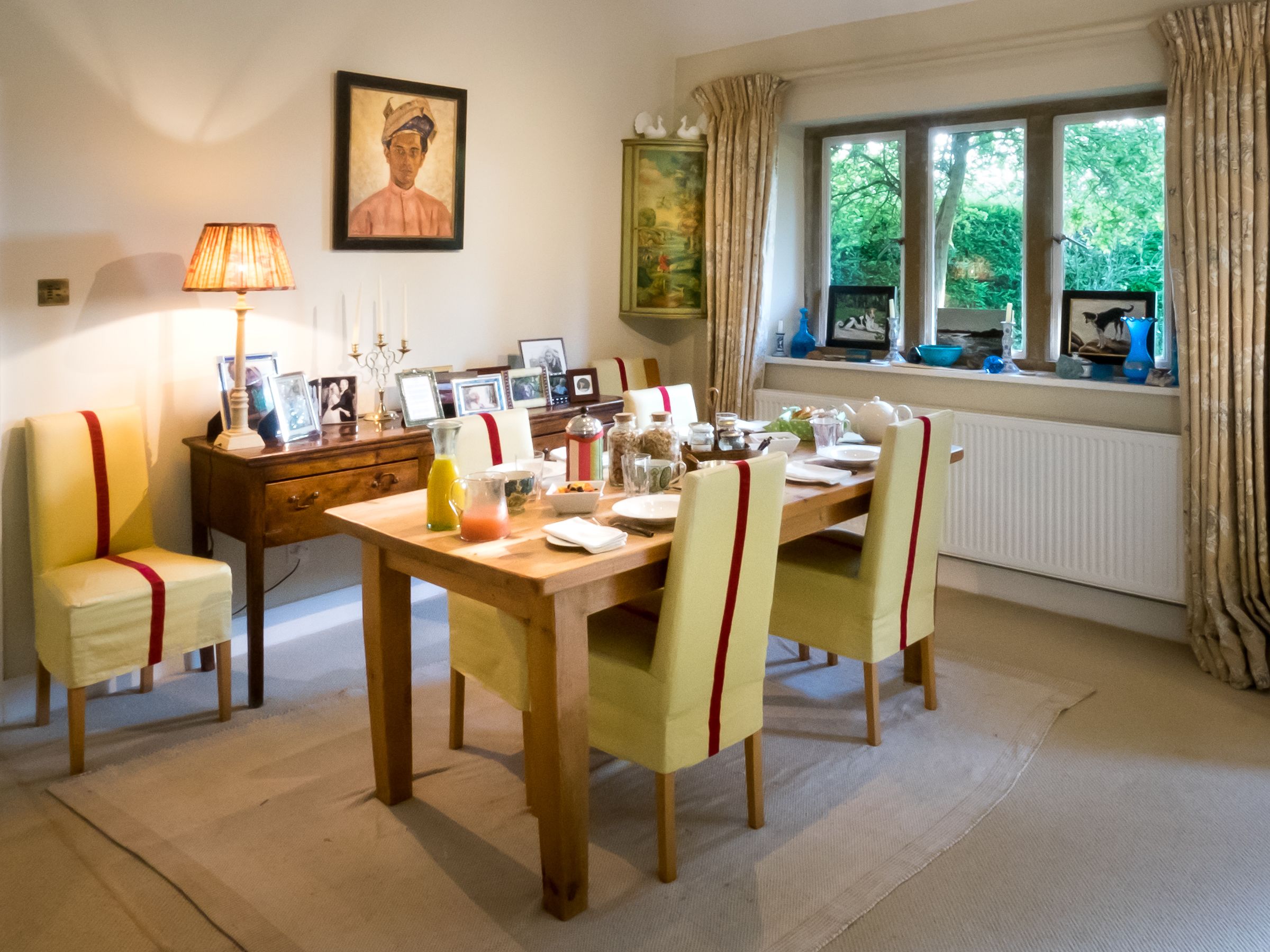 Een eetkamer met een houten tafel gedekt voor vier personen, omringd door stoelen met rode linten. Een lamp, een schilderij en een raam met gordijnen zijn zichtbaar.