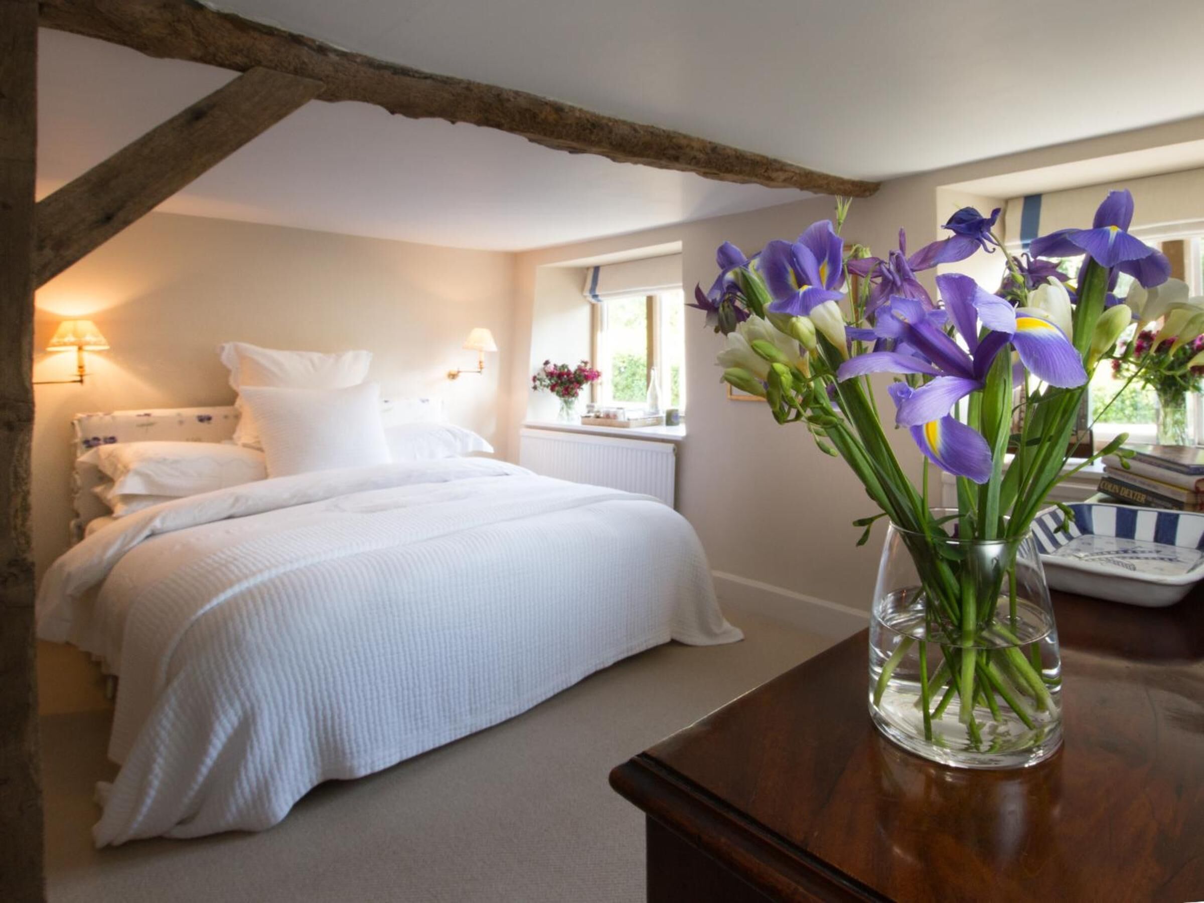 A bedroom with a large bed, a wooden ceiling beam, a bedside table with a vase of flowers, and a window with a view of greenery.