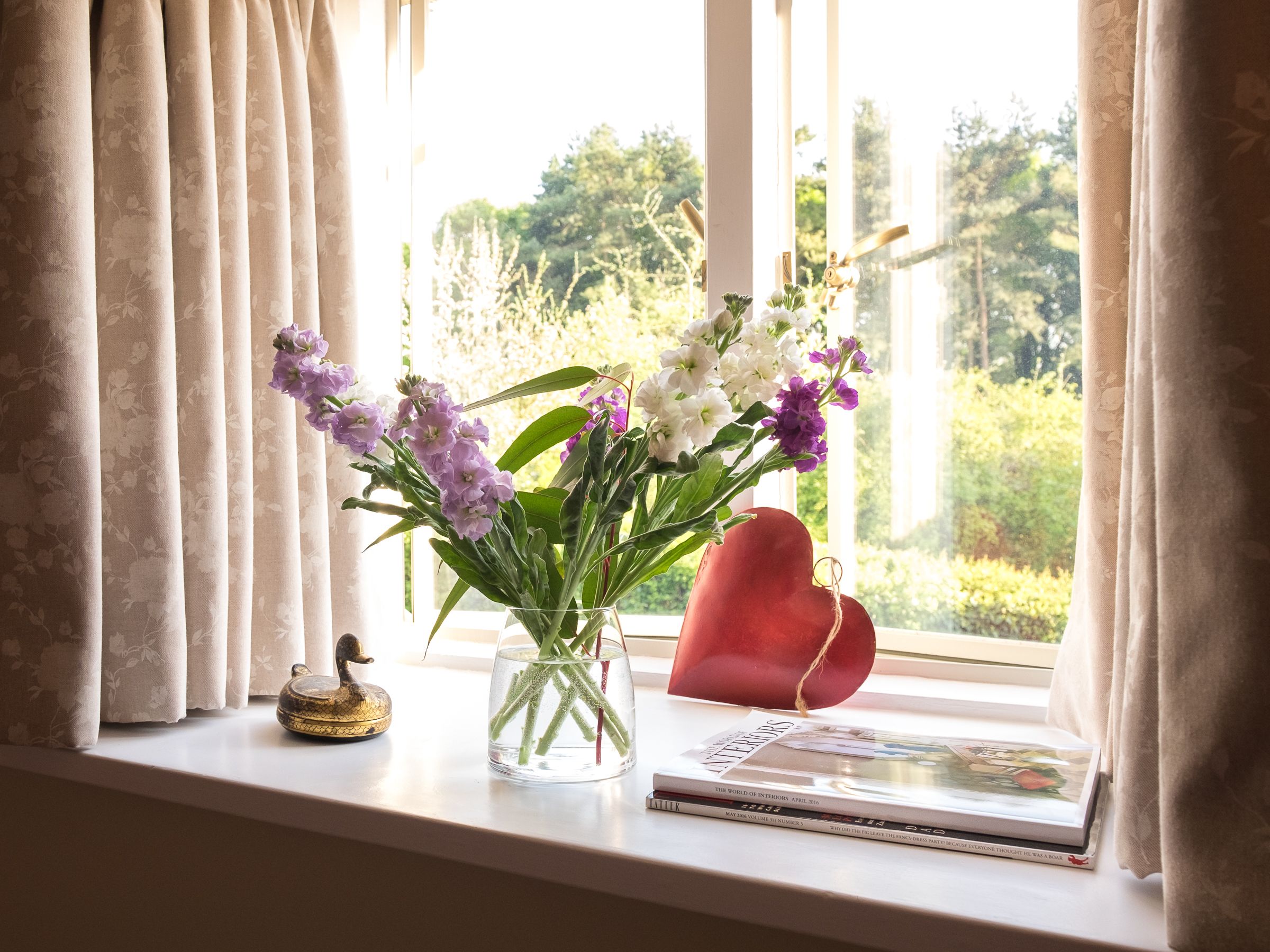 A window with a vase of flowers, a heart-shaped decoration, and a book on a windowsill.