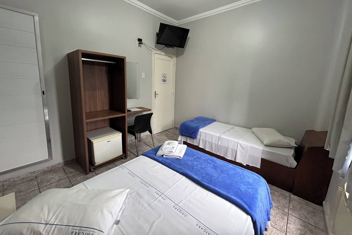 Quarto de hotel com duas camas, uma escrivaninha, um guarda-roupa e uma televisão na parede.