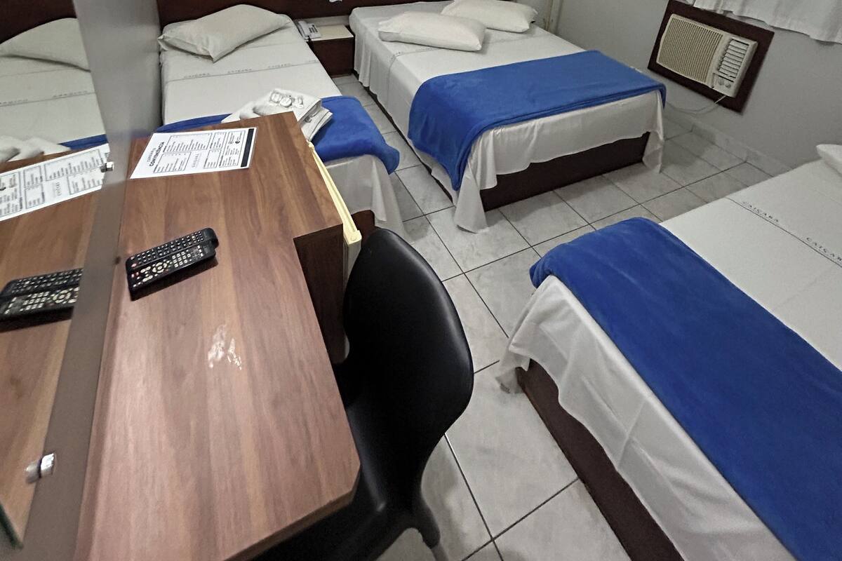 Quarto de hotel com três camas de solteiro, uma escrivaninha de madeira com cadeira, mesa de cabeceira e uma janela com cortinas.