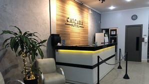 A reception desk with a wooden panel and a sign, a waiting area with chairs, a potted plant, and a security chain. - Hotel Caiçara (Santos)