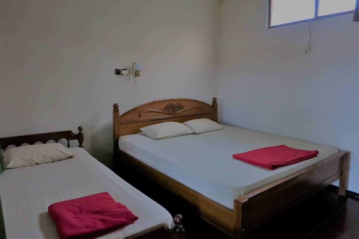 A room with two single beds, each with a wooden headboard and white bedding. There is a window with blinds on the wall.