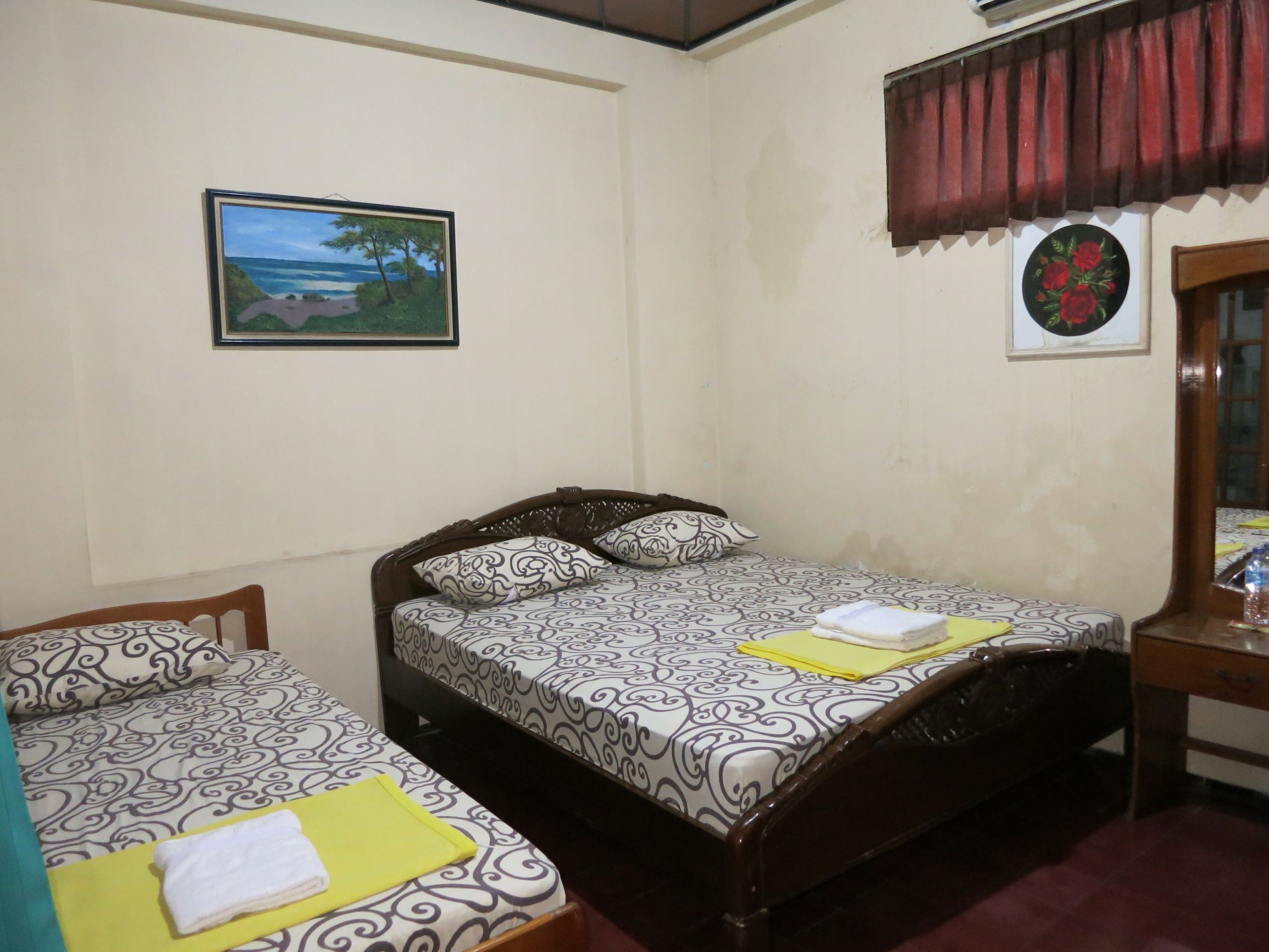 Ein Schlafzimmer mit zwei Betten, einem Strandbild, einem Fenster mit rotem Vorhang und einem kleinen Tisch mit Spiegel.